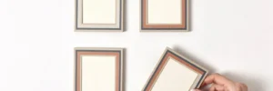 Vier identieke kleine lijstjes in een strak grid op wit oppervlak, één frame voorzichtig bijgesteld door een hand, in warme neutrale tinten met leisteen blauw en terracotta.